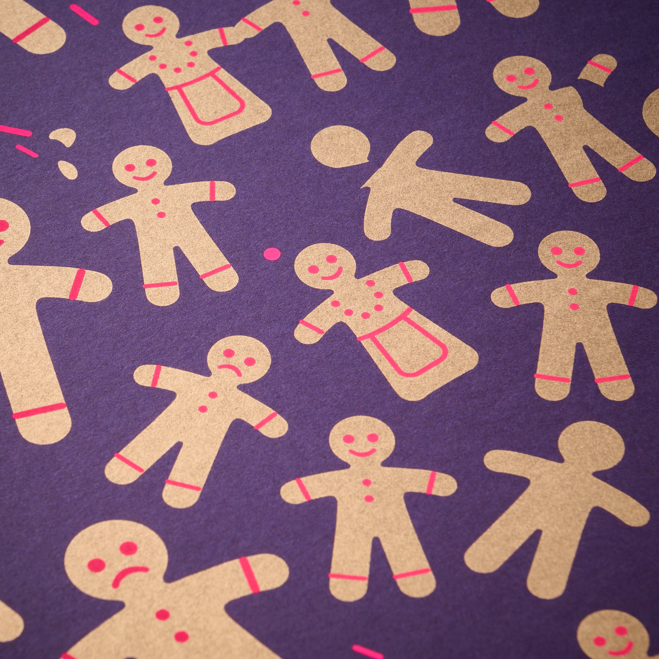 Wrapping paper with a festive pattern of golden gingerbread men and women scattered across a deep purple background, accented with small bright pink details.