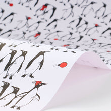 Wrapping paper featuring a lively crowd of black-and-white penguins against a white background, with scattered red holiday accents such as ornaments and hats adding a cheerful pop of color.