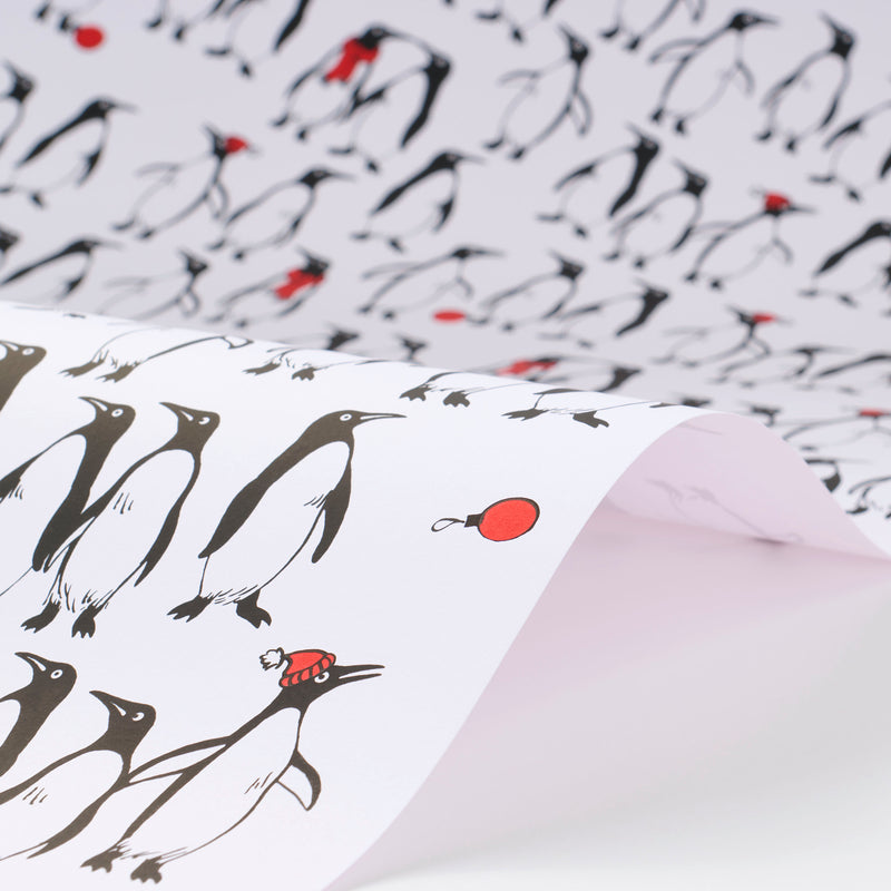 Wrapping paper featuring a lively crowd of black-and-white penguins against a white background, with scattered red holiday accents such as ornaments and hats adding a cheerful pop of color.