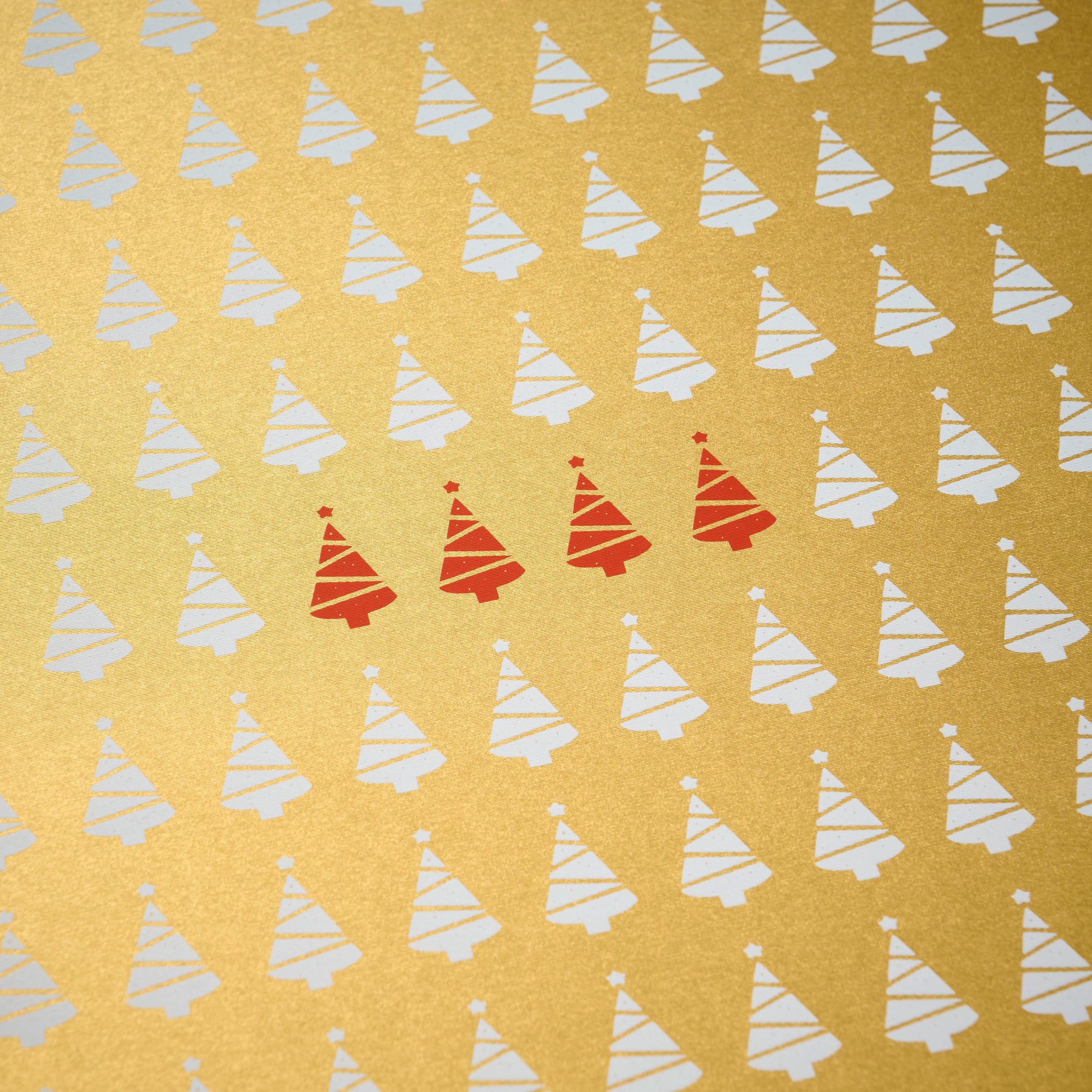 Wrapping paper with a festive pattern of tiny white Christmas trees scattered across a shimmering gold background, accented by a small row of bright red trees.