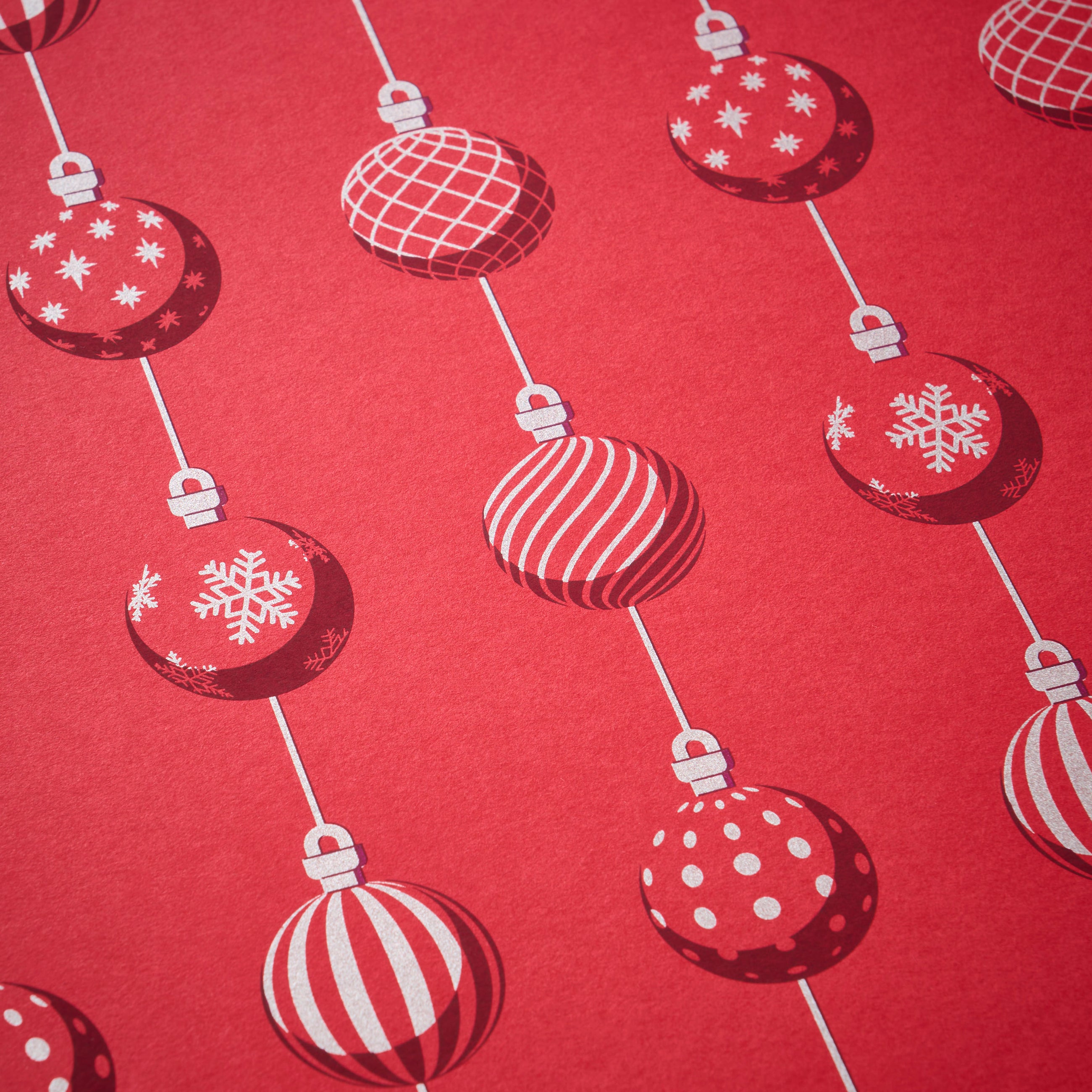 Wrapping paper with a festive pattern of red Christmas baubles decorated in white snowflakes, stripes, grids, and polka dots, arranged in vertical lines against a bright red background.
