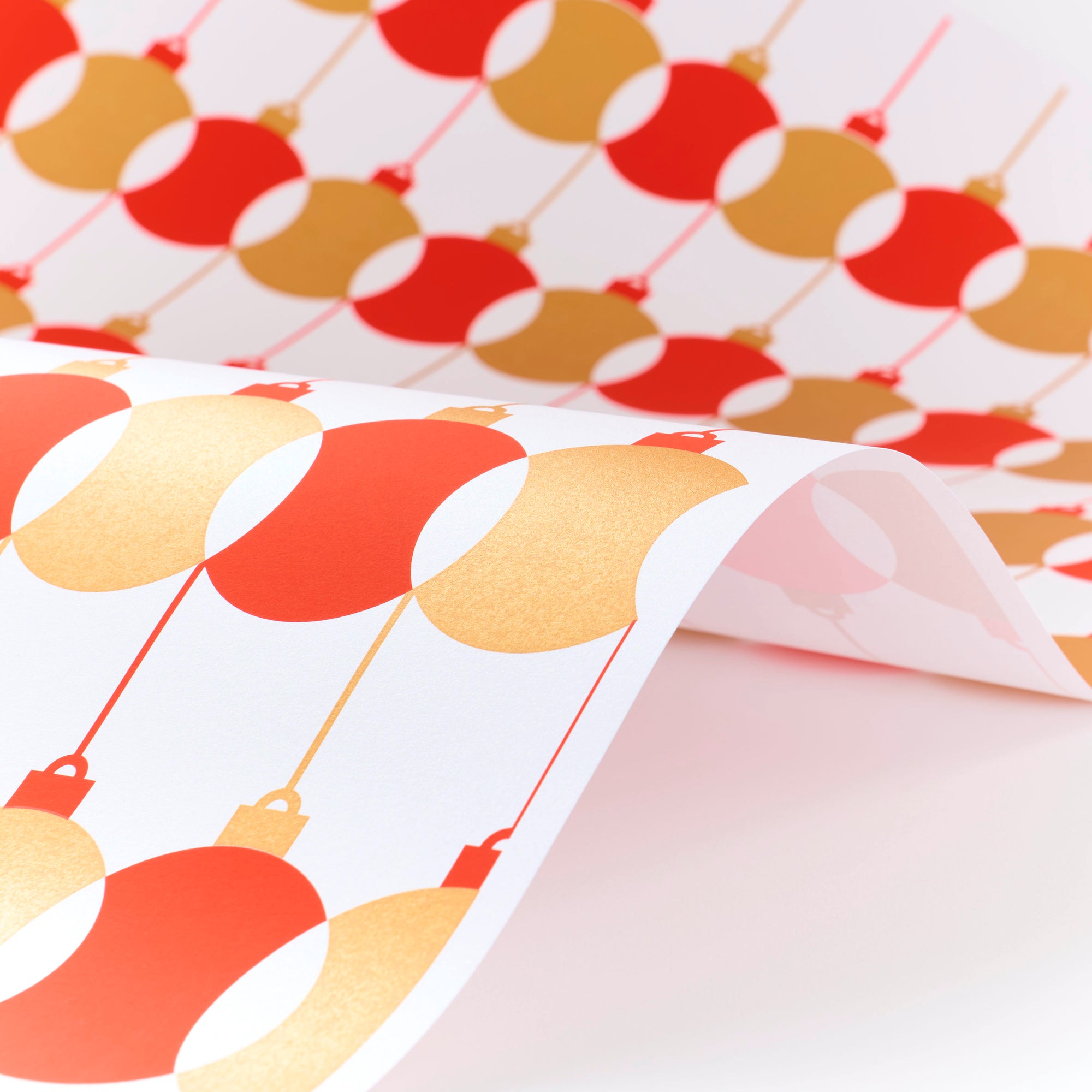 Wrapping paper with a festive pattern of red and gold baubles repeating in neat rows, shown curled to reveal its crisp white background.