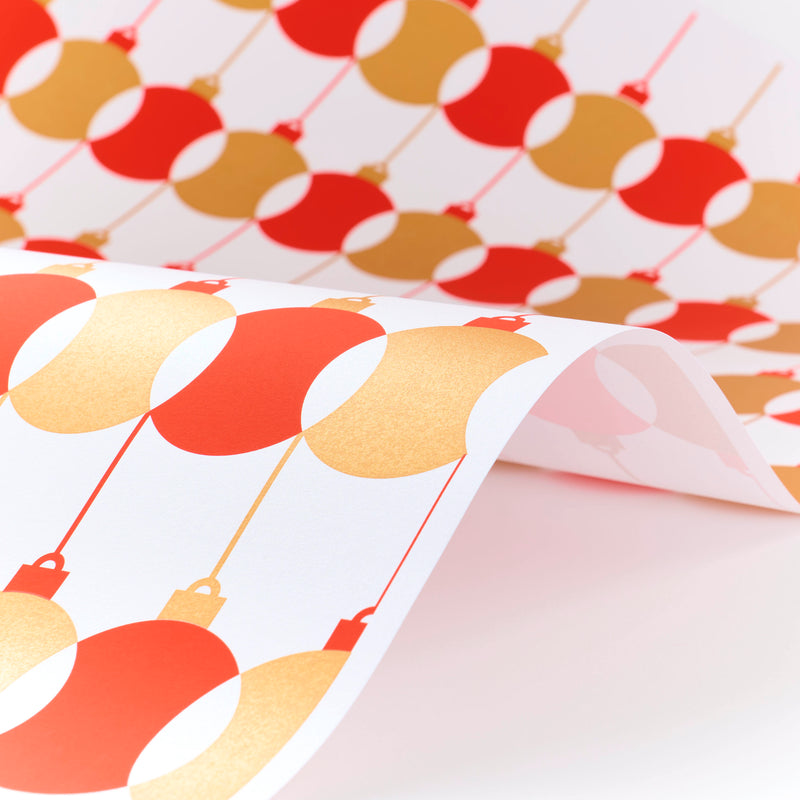 Wrapping paper with a festive pattern of red and gold baubles repeating in neat rows, shown curled to reveal its crisp white background.