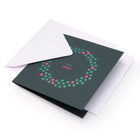 A dark green Christmas card with a minimalist wreath design made of green and pink dots, surrounding the text "VERY MERRY" in pink and white at the center, shown with a plain white envelope.