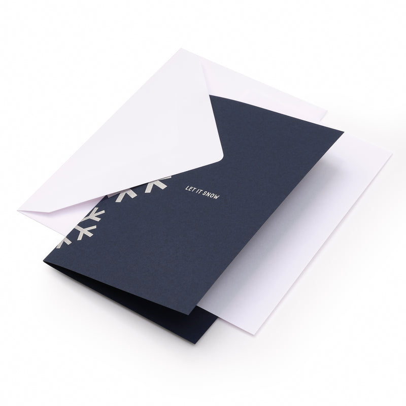 A dark blue holiday card with silver geometric snowflakes along the left edge and the text "LET IT SNOW" in silver at the center, shown with a plain white envelope.