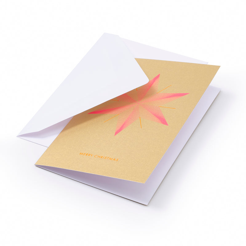 A gold Christmas card with a bold pink and orange geometric star design in the center and the text "MERRY CHRISTMAS" in orange at the bottom, shown with a plain white envelope.
