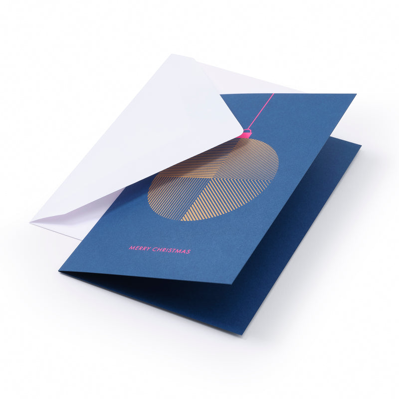 A dark blue Christmas card with a minimalist gold ornament design featuring fine geometric lines, a pink string and hook, and the text "MERRY CHRISTMAS" in pink at the bottom, shown with a plain white envelope.