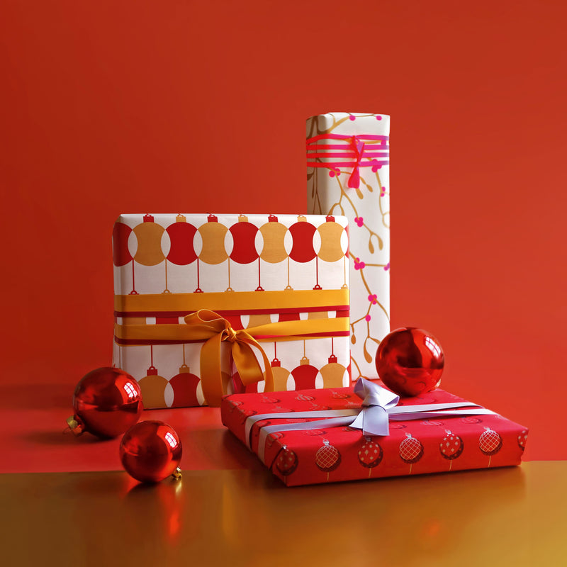 Three Christmas gifts wrapped in festive paper with red, gold, and white designs featuring ornament and branch patterns, tied with coordinating ribbons, surrounded by shiny red baubles on a red and gold background.
