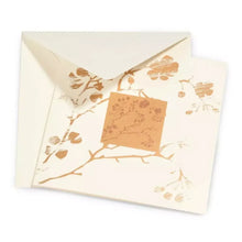 A refined greeting card with a soft cream background, adorned with delicate, metallic gold botanical prints of branches, leaves, and cherry blossoms arranged around a central square motif, partially tucked into a matching envelope.