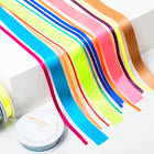 A set of colorful satin ribbons in bright neon and metallic shades, including pink, blue, green, yellow, orange, purple, and gold, arranged in parallel strips with ribbon spools placed beside them.