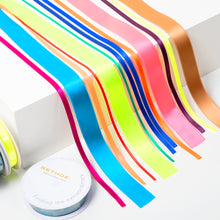 A set of colorful satin ribbons in bright neon and metallic shades, including pink, blue, green, yellow, orange, purple, and gold, arranged in parallel strips with ribbon spools placed beside them.