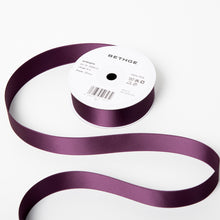 A spool of satin ribbon in deep aubergine purple, partially unrolled, with a white label on top reading “BETHGE” and product details including width, length, and material.