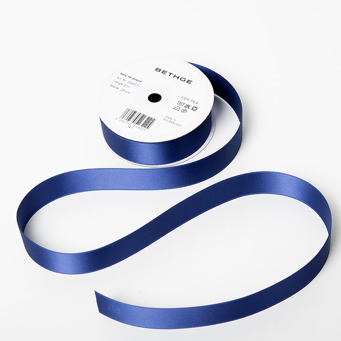 A spool of satin ribbon in plum blue, partially unrolled, with a white label on top reading “BETHGE” and product details including width, length, and material.