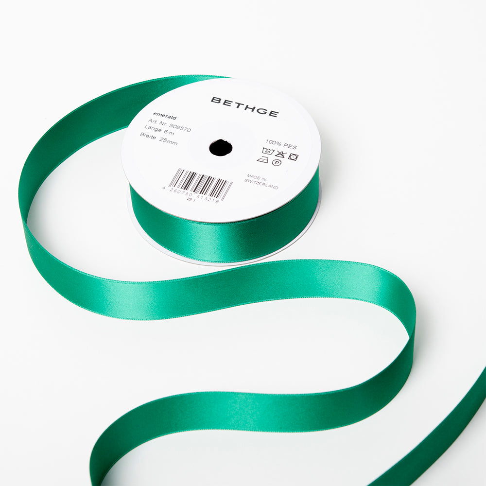A spool of satin ribbon in emerald green, partially unrolled, with a white label on top reading “BETHGE” and product details including width, length, and material.