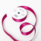 A spool of satin ribbon in fuchsia pink, partially unrolled, with a white label on top reading “BETHGE” and product details including width, length, and material.