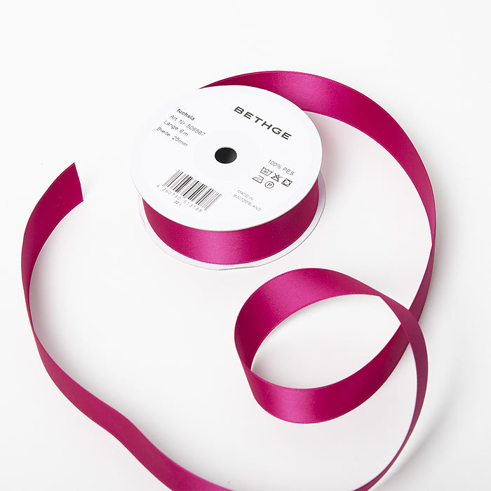A spool of satin ribbon in fuchsia pink, partially unrolled, with a white label on top reading “BETHGE” and product details including width, length, and material.