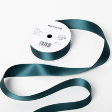 A spool of satin ribbon in hunter green, partially unrolled, with a white label on top reading “BETHGE” and product details including width, length, and material.