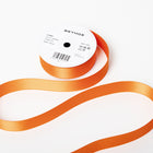 A spool of satin ribbon in mango orange, partially unrolled, with a white label on top reading “BETHGE” and product details including width, length, and material.