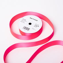 A spool of satin ribbon in neon pink, partially unrolled, with a white label on top reading “BETHGE” and product details including width, length, and material.