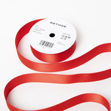 A spool of satin ribbon in bright red, partially unrolled, with a white label on top reading “BETHGE” and product details including width, length, and material.