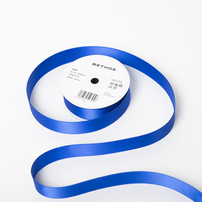 A spool of satin ribbon in royal blue, partially unrolled, with a white label on top reading “BETHGE” and product details including width, length, and material.