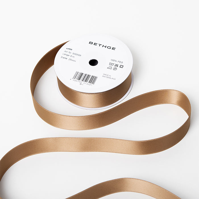 A spool of satin ribbon in sable brown, partially unrolled, with a white label on top reading “BETHGE” and product details including width, length, and material.