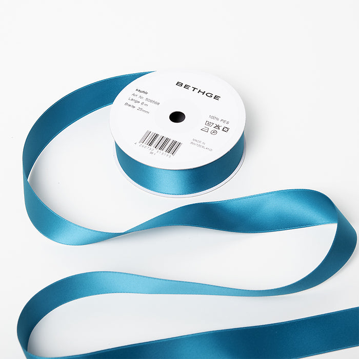 A spool of satin ribbon in sapphire blue, partially unrolled, with a white label on top reading “BETHGE” and product details including width, length, and material.