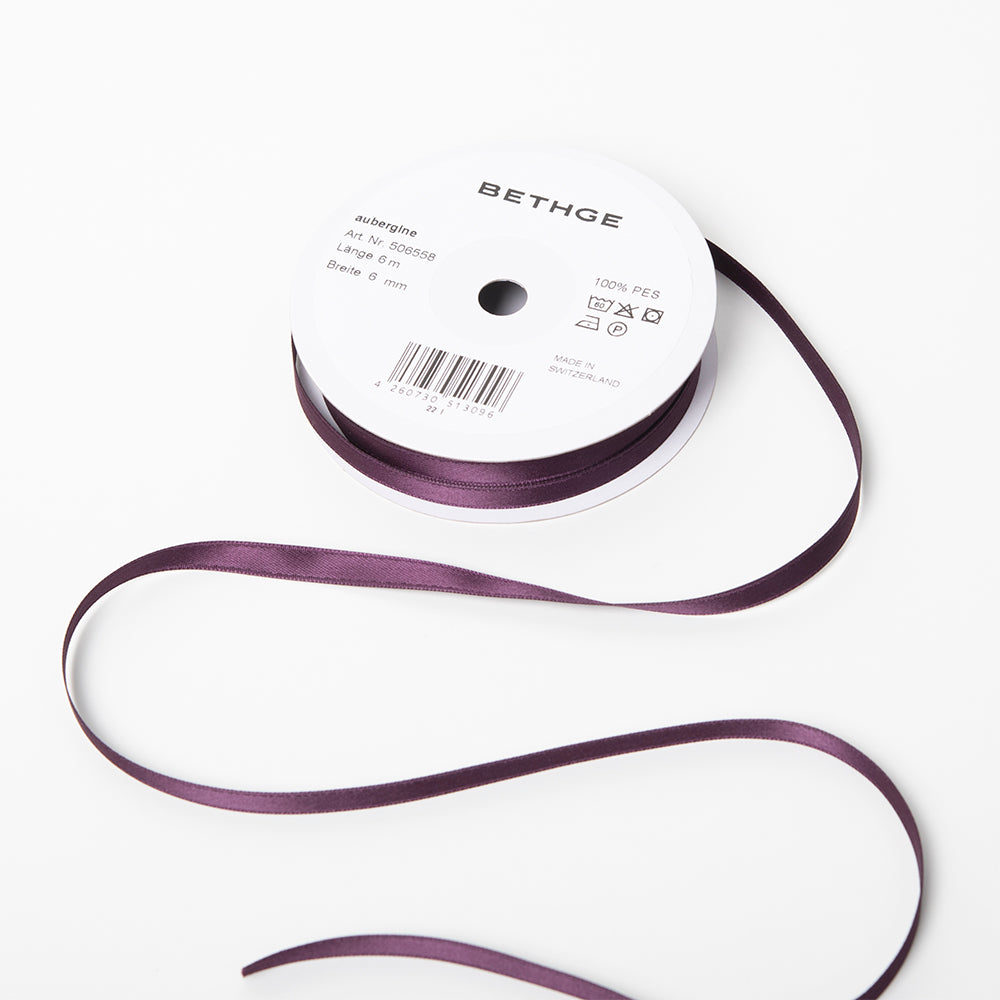 A spool of satin ribbon in deep aubergine purple, narrow in width, partially unrolled, with a white label on top reading “BETHGE” and product details including width, length, and material.