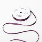 A spool of satin ribbon in deep aubergine purple, narrow in width, partially unrolled, with a white label on top reading “BETHGE” and product details including width, length, and material.