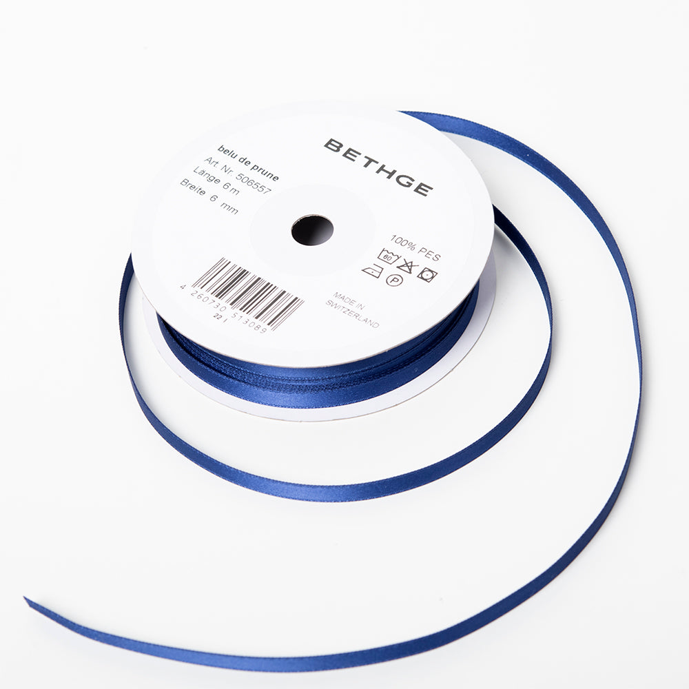 A spool of satin ribbon in plum blue, narrow in width, partially unrolled, with a white label on top reading “BETHGE” and product details including width, length, and material.