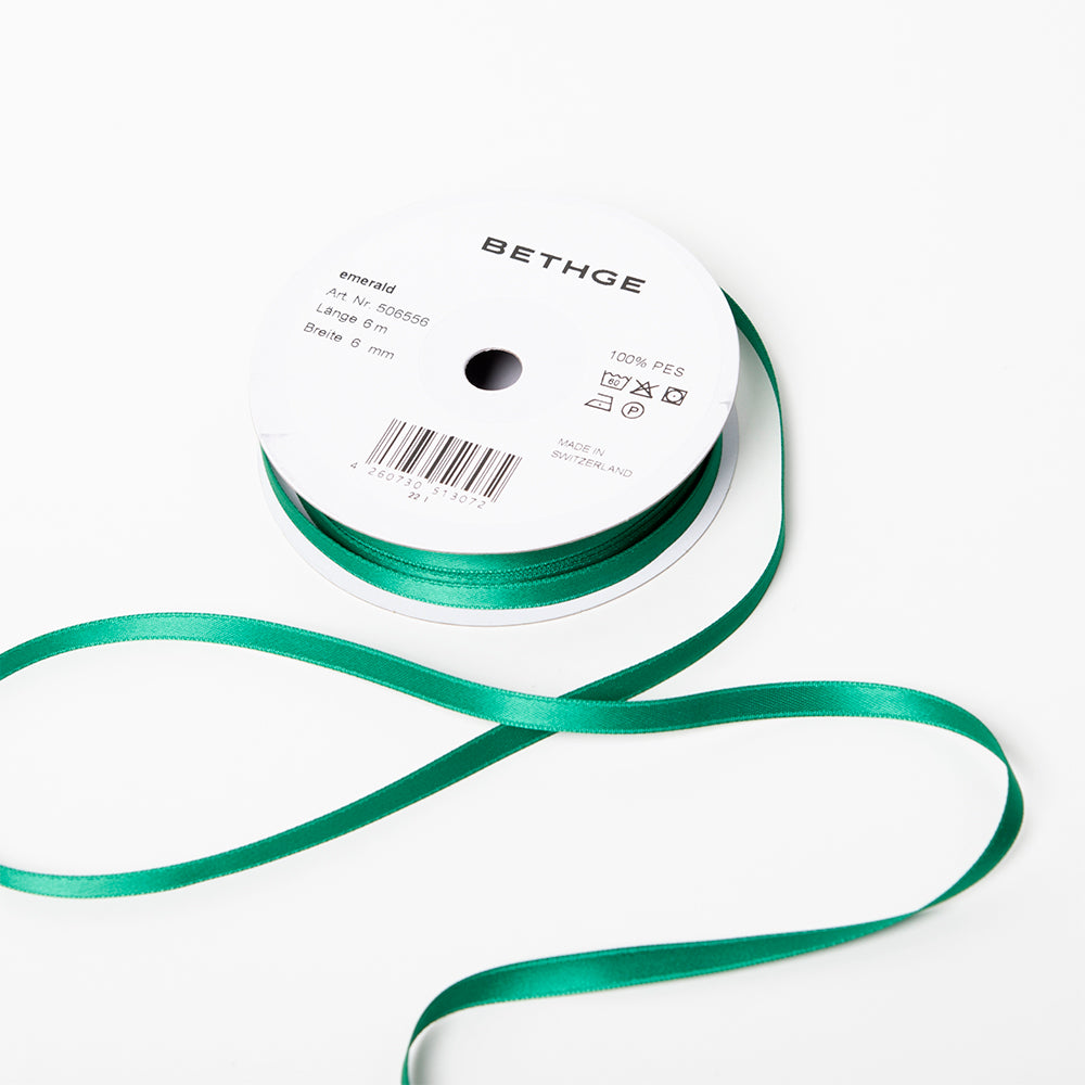A spool of satin ribbon in emerald green, narrow in width, partially unrolled, with a white label on top reading “BETHGE” and product details including width, length, and material.