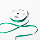 A spool of satin ribbon in emerald green, narrow in width, partially unrolled, with a white label on top reading “BETHGE” and product details including width, length, and material.