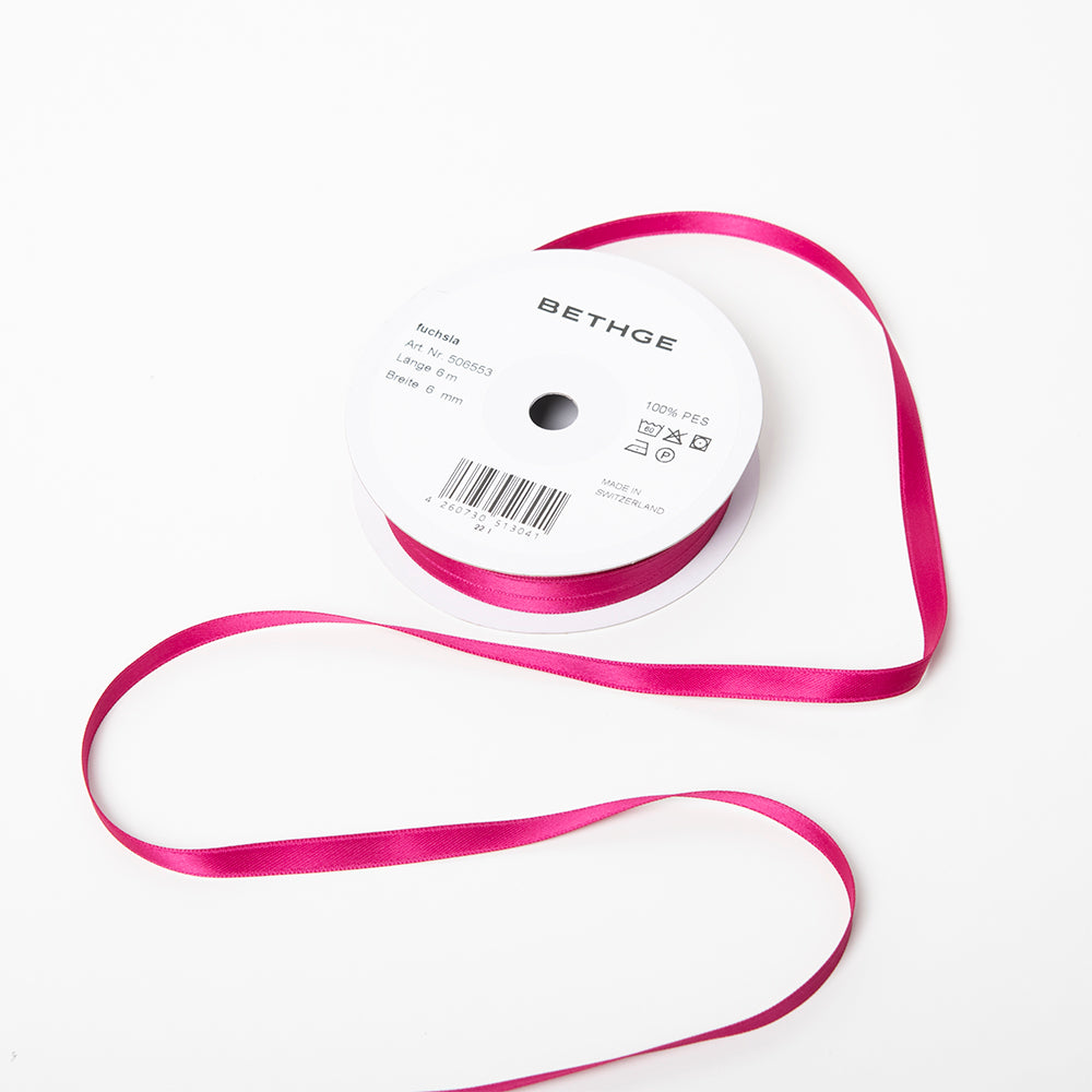 A spool of satin ribbon in fuchsia pink, narrow in width, partially unrolled, with a white label on top reading “BETHGE” and product details including width, length, and material.