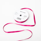 A spool of satin ribbon in fuchsia pink, narrow in width, partially unrolled, with a white label on top reading “BETHGE” and product details including width, length, and material.