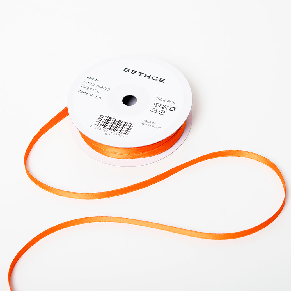 A spool of satin ribbon in mango orange, narrow in width, partially unrolled, with a white label on top reading “BETHGE” and product details including width, length, and material.