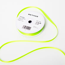 A spool of satin ribbon in neon green, narrow in width, partially unrolled, with a white label on top reading “BETHGE” and product details including width, length, and material.