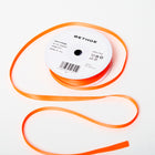 A spool of satin ribbon in neon orange, narrow in width, partially unrolled, with a white label on top reading “BETHGE” and product details including width, length, and material.