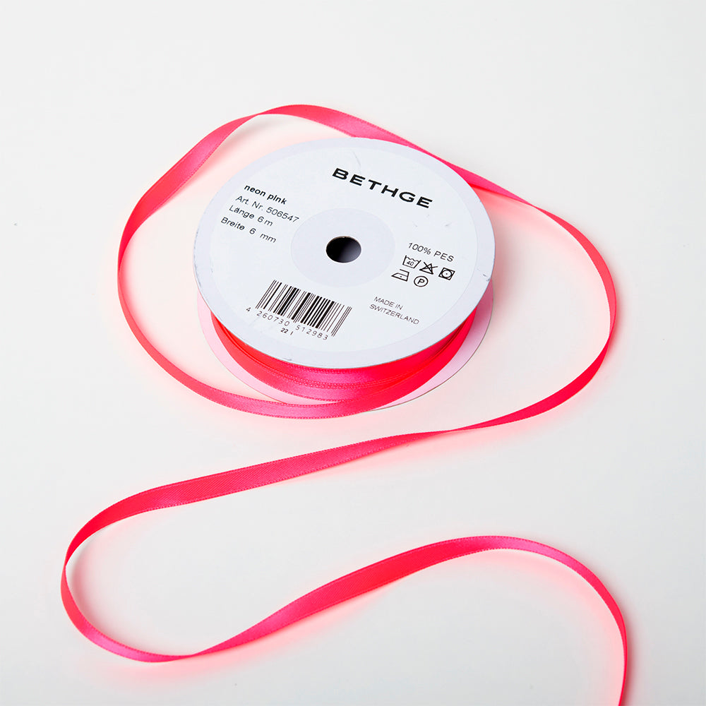 A spool of satin ribbon in neon pink, narrow in width, partially unrolled, with a white label on top reading “BETHGE” and product details including width, length, and material.