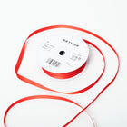A spool of satin ribbon in bright red, narrow in width, partially unrolled, with a white label on top reading “BETHGE” and product details including width, length, and material.
