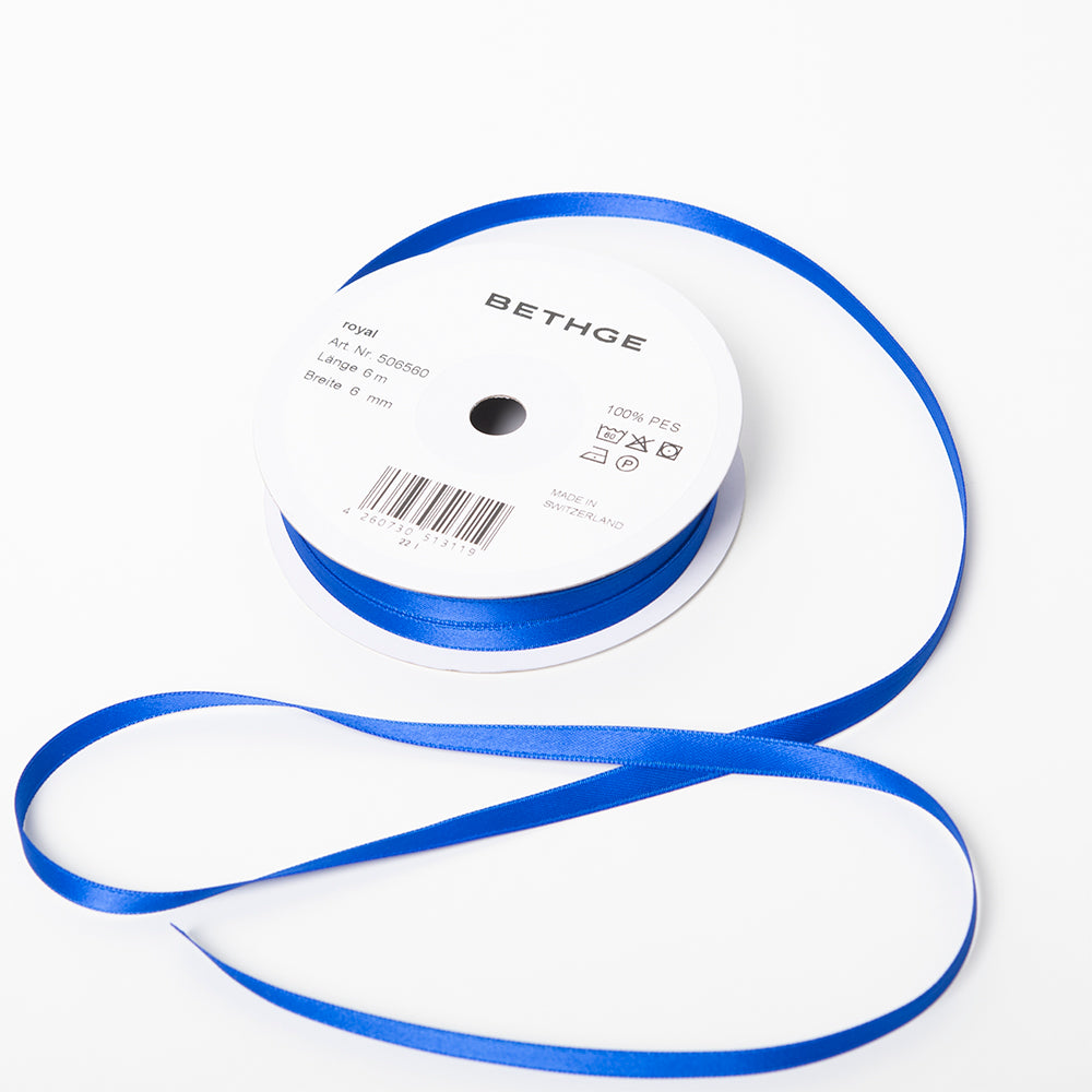 A spool of satin ribbon in royal blue, narrow in width, partially unrolled, with a white label on top reading “BETHGE” and product details including width, length, and material.