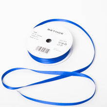 A spool of satin ribbon in royal blue, narrow in width, partially unrolled, with a white label on top reading “BETHGE” and product details including width, length, and material.