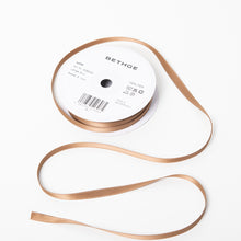 A spool of satin ribbon in sable brown, narrow in width, partially unrolled, with a white label on top reading “BETHGE” and product details including width, length, and material.