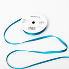 A spool of satin ribbon in sapphire blue, narrow in width, partially unrolled, with a white label on top reading “BETHGE” and product details including width, length, and material.