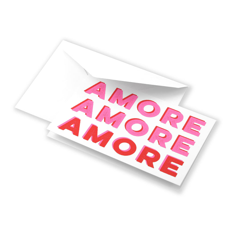 Greeting card partially pulled from a white envelope, featuring the word “AMORE” repeated in large pink and red letters, creating a playful, modern love-themed design.