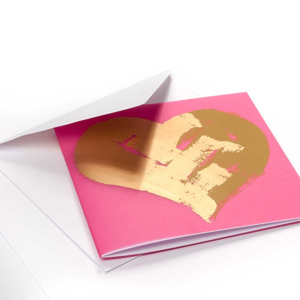 Greeting card with a vibrant pink background, featuring a large hand-painted gold heart motif with expressive brush strokes, shown partially tucked into a white envelope.