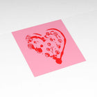 Greeting card with a minimalist design, featuring a hand-stamped red heart with dotted texture on a soft pink square background, photographed flat on a white surface.