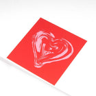 Greeting card with a minimalist design, featuring a painterly light-pink heart printed on a bold red square background, shown at a slight angle against a clean white surface.