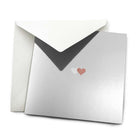 Greeting card with a minimalist design, featuring a reflective silver surface and two small overlapping hearts, one soft white, one warm bronze, centered on the card, partially tucked into a matching white envelope.