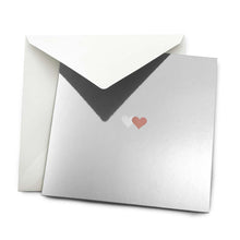Greeting card with a minimalist design, featuring a reflective silver surface and two small overlapping hearts, one soft white, one warm bronze, centered on the card, partially tucked into a matching white envelope.