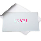 Silver greeting card with a metallic finish and the word “LOVE!” cut out in bright pink, shown with a white envelope behind it.